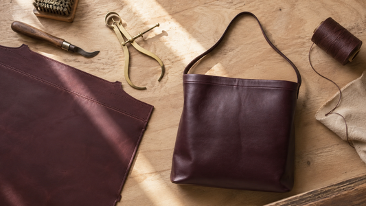 Flat lay of a burgundy leather tote bag alongside brass calipers, a cutting knife, and waxed thread on a wooden workbench.